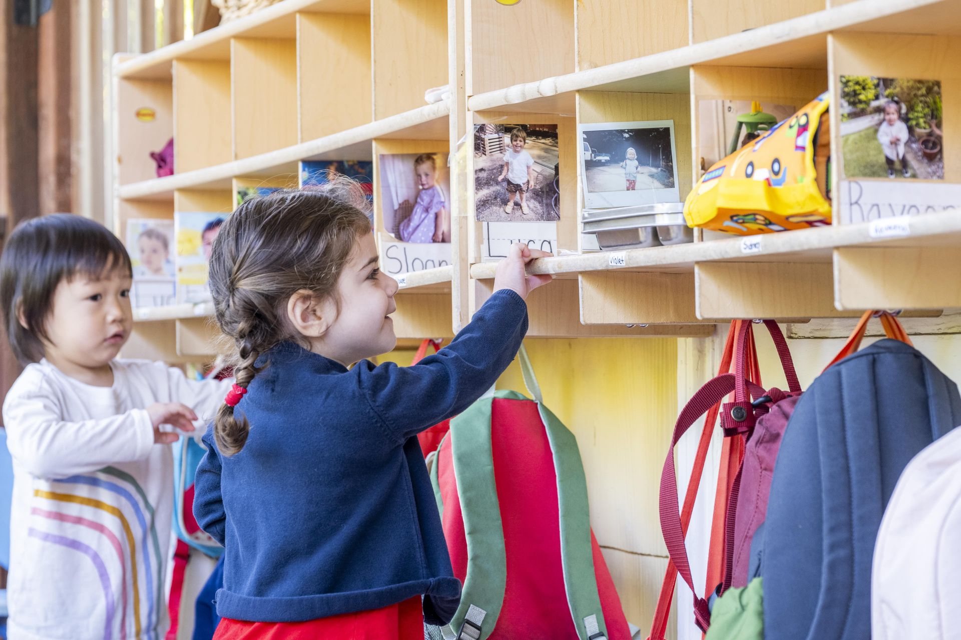 A Montessori child is reaching for a backpack on a shelf.