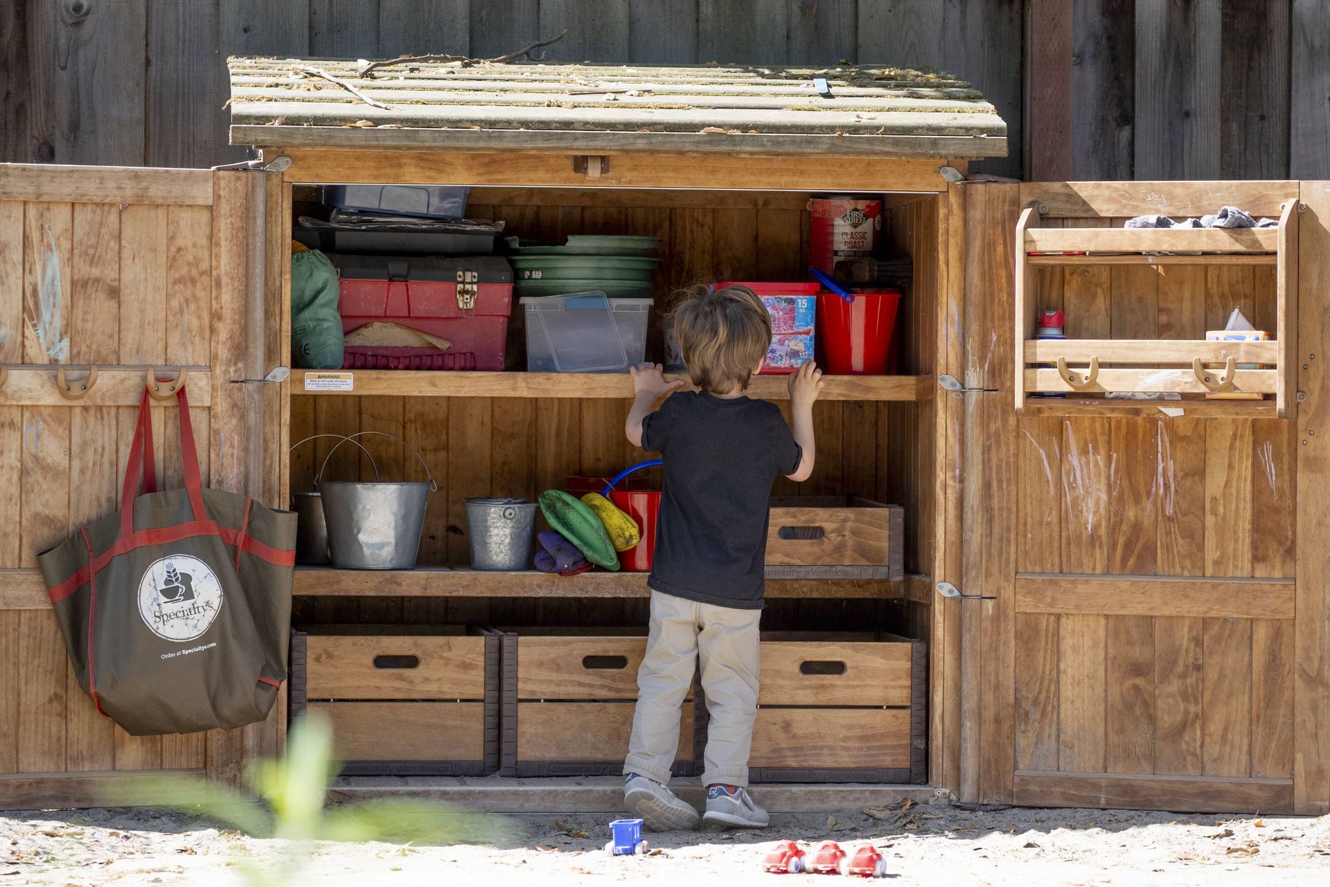 A Montessori child is standing in front of a wooden shed filled with toys.