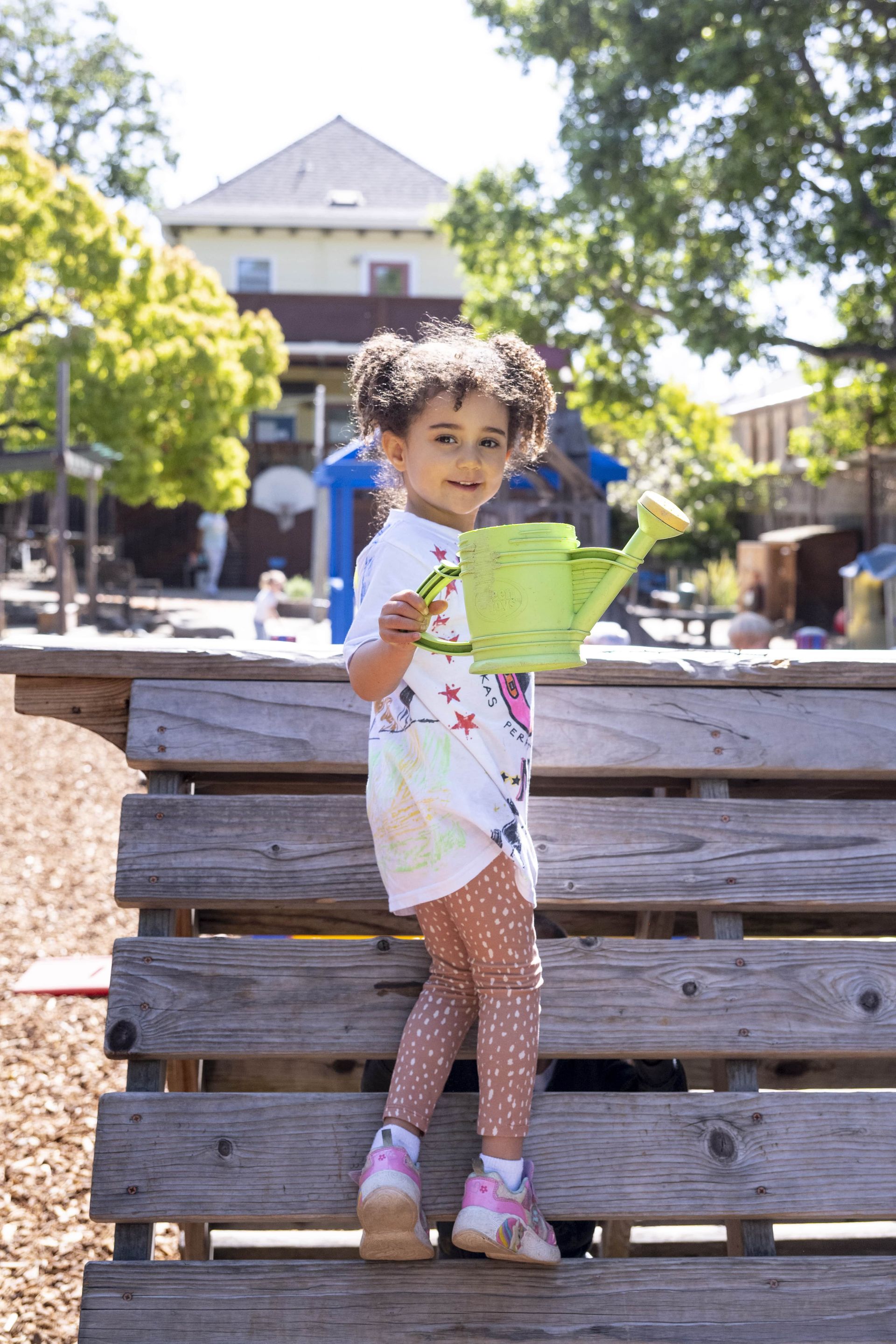 Montessori child is standing on a wooden bench holding a watering can.