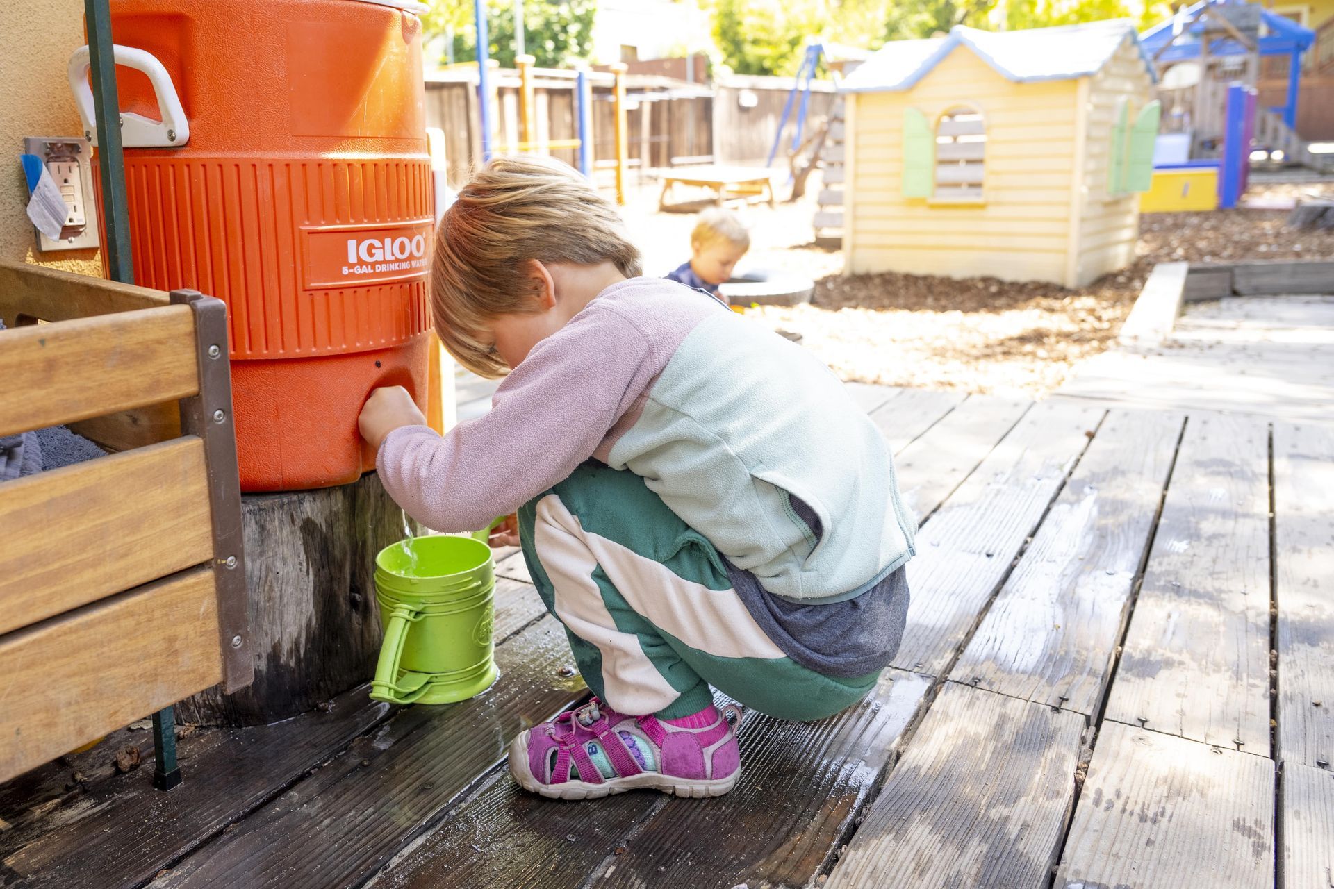 A Montessori child is kneeling down on a wooden deck holding a green bucket.