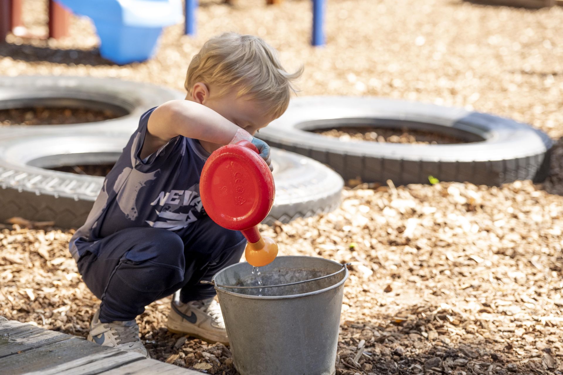 A Montessori child is pouring water from a red watering can into a bucket.