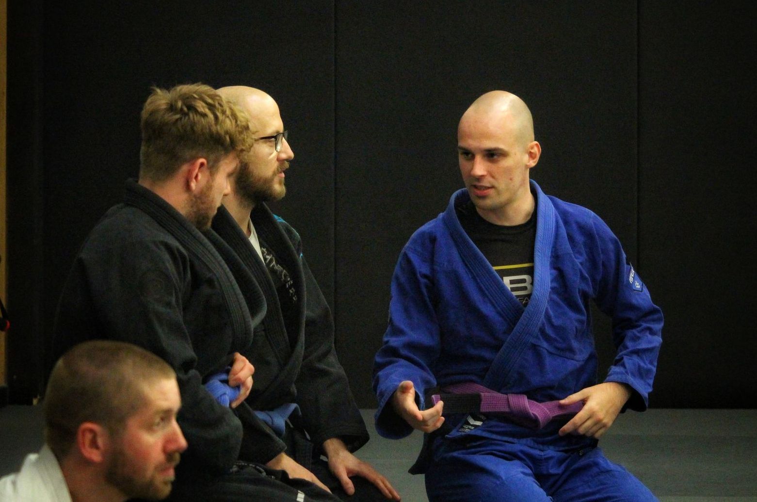 Three people in martial arts uniforms sit talking in a dim studio setting