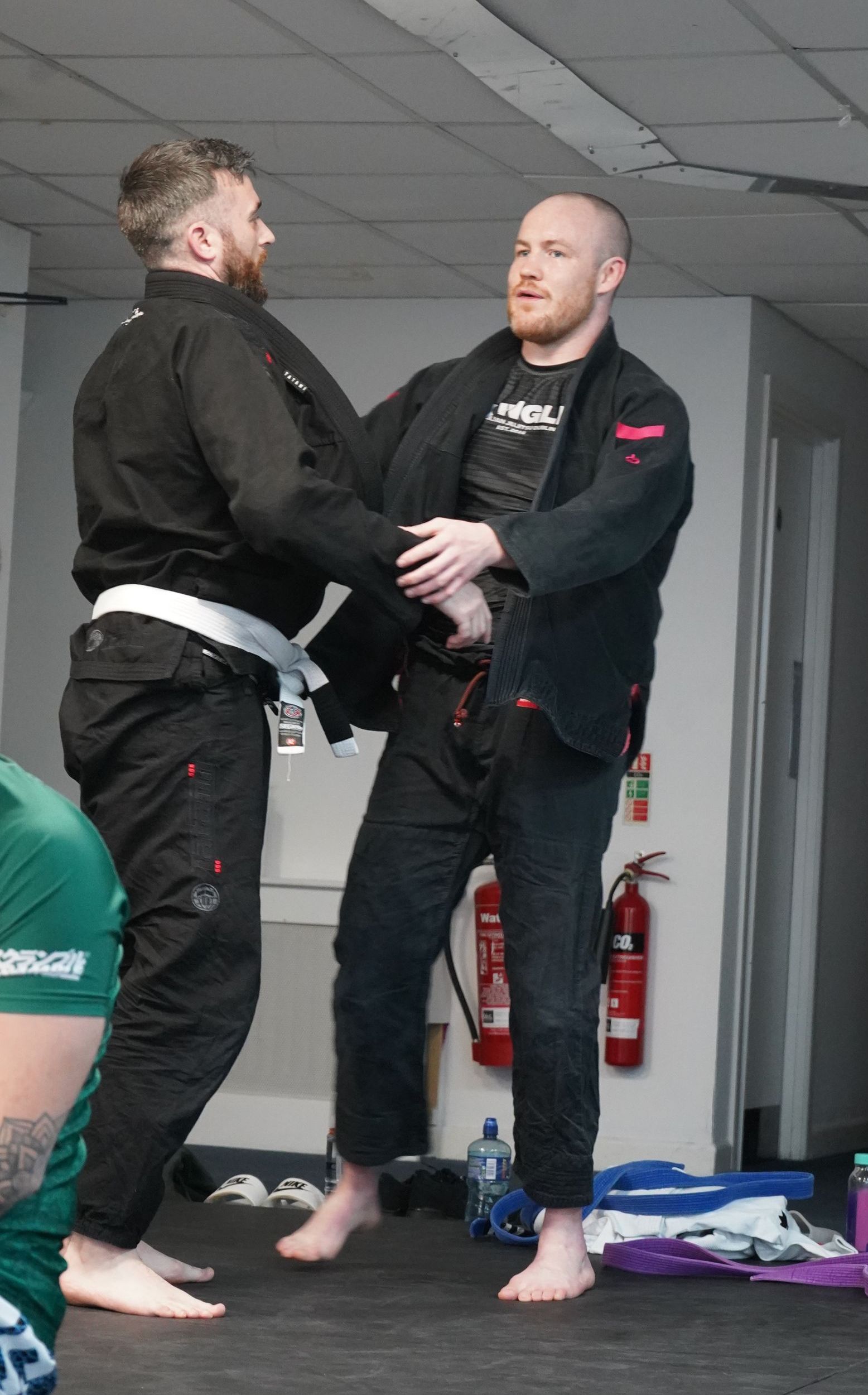 Two martial artists in black gis practice a grip drill indoors on mats.