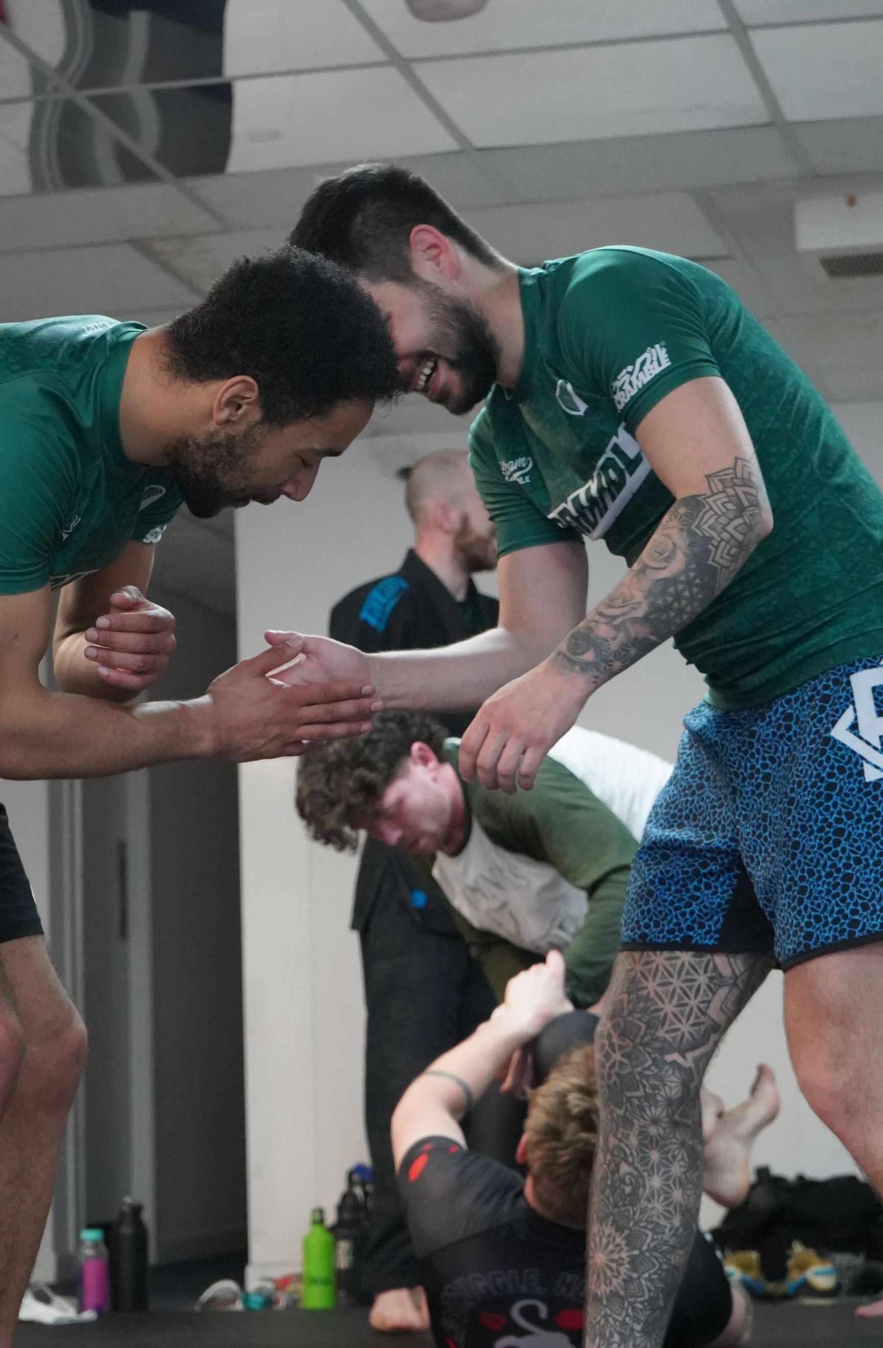 Two athletes in green jerseys kneel and clasp hands in a locker room, with another person seated behind them.