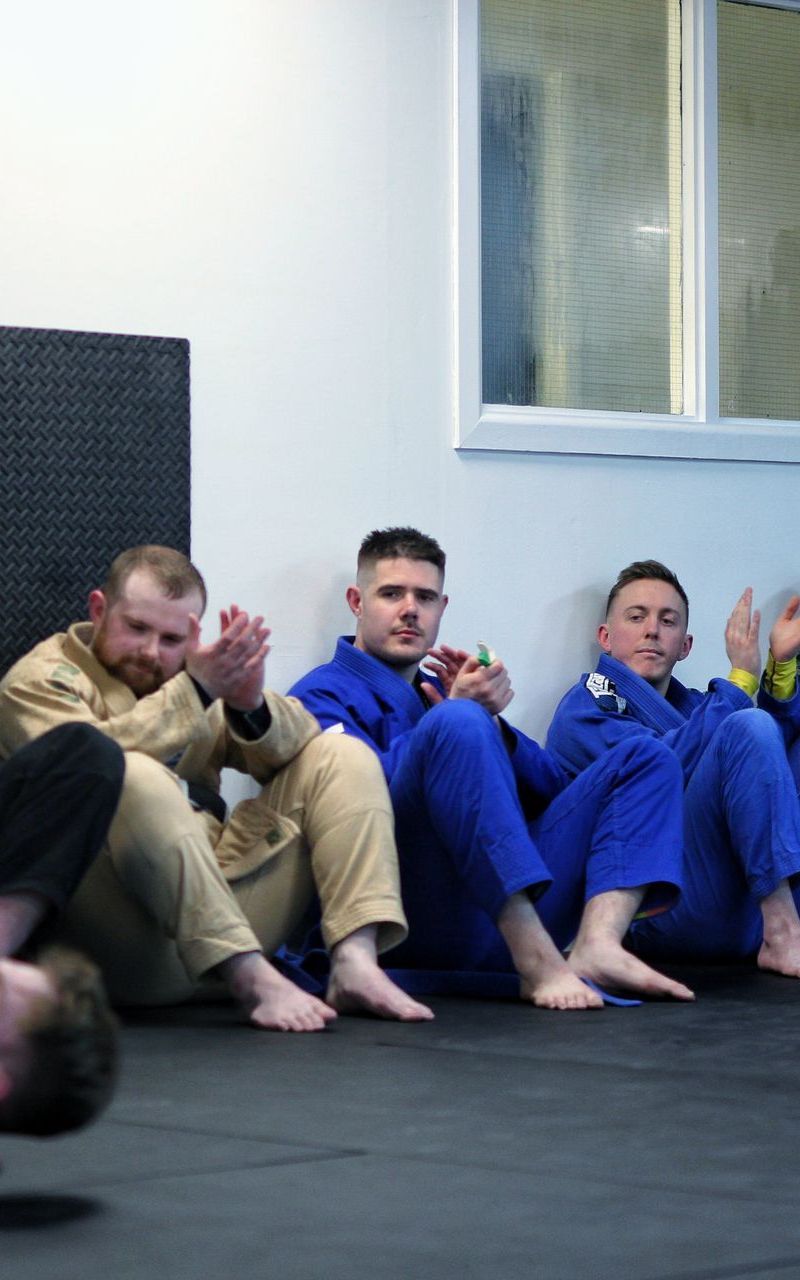 Four people in blue and tan gis sit on a mat against a wall, relaxing in a martial arts studio.