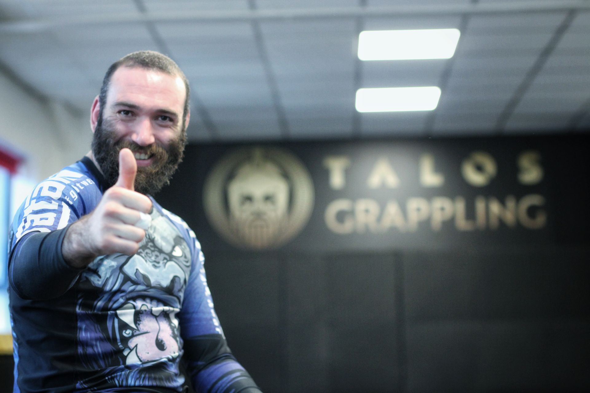 Man in a blue rash guard giving a thumbs-up in a Talos Grappling gym