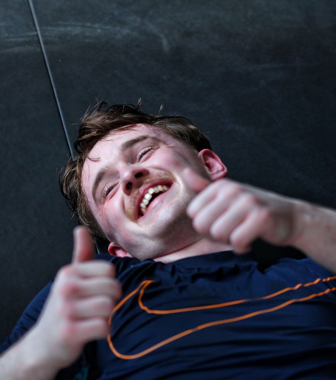 Smiling man giving two thumbs up while lying on a dark floor in a navy shirt