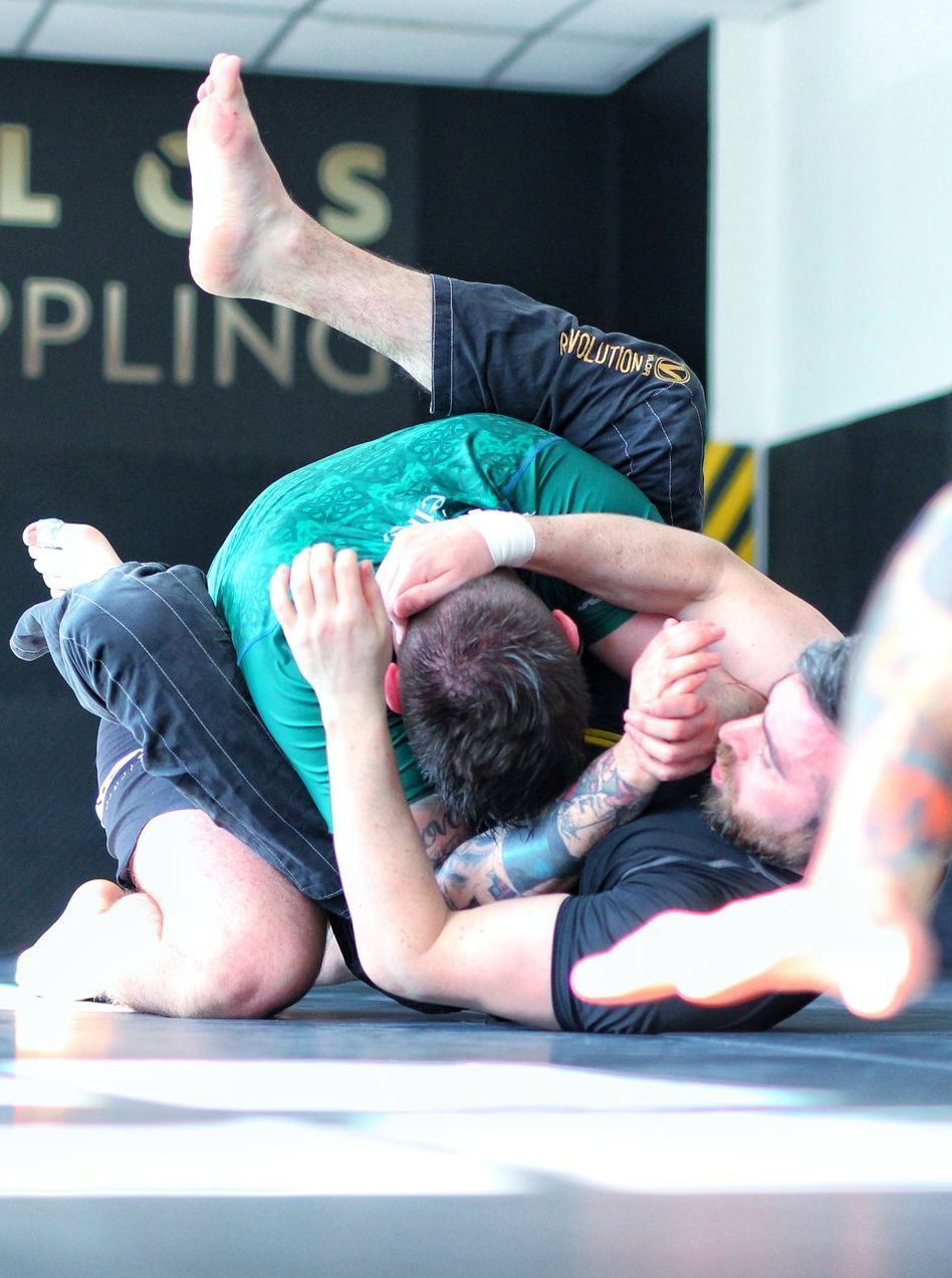 Two wrestlers grappling on a mat, one in a teal shirt on top, with another athlete in black.