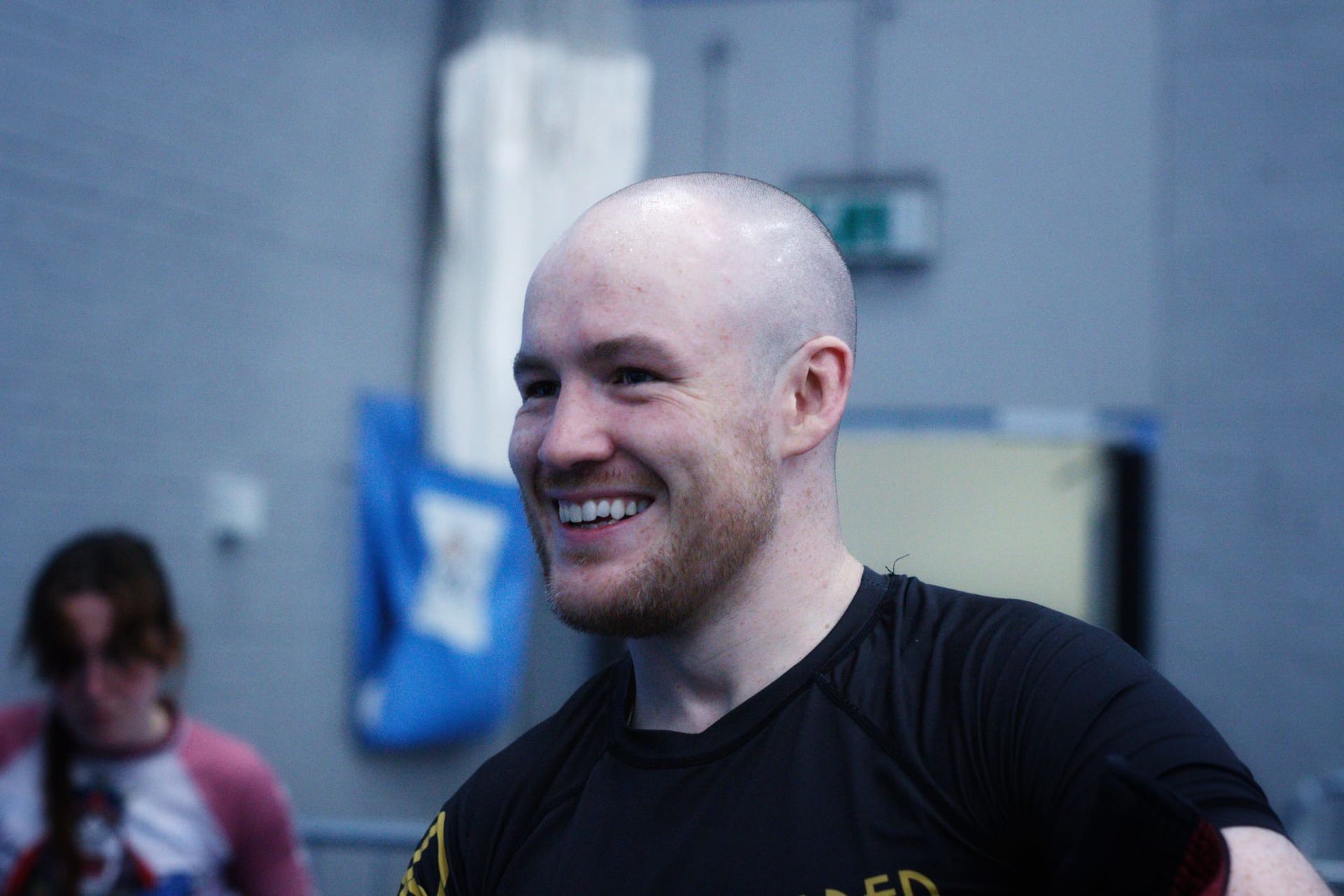 Smiling bald man in a black shirt at an indoor event, with a blurred person and blue bag in the background
