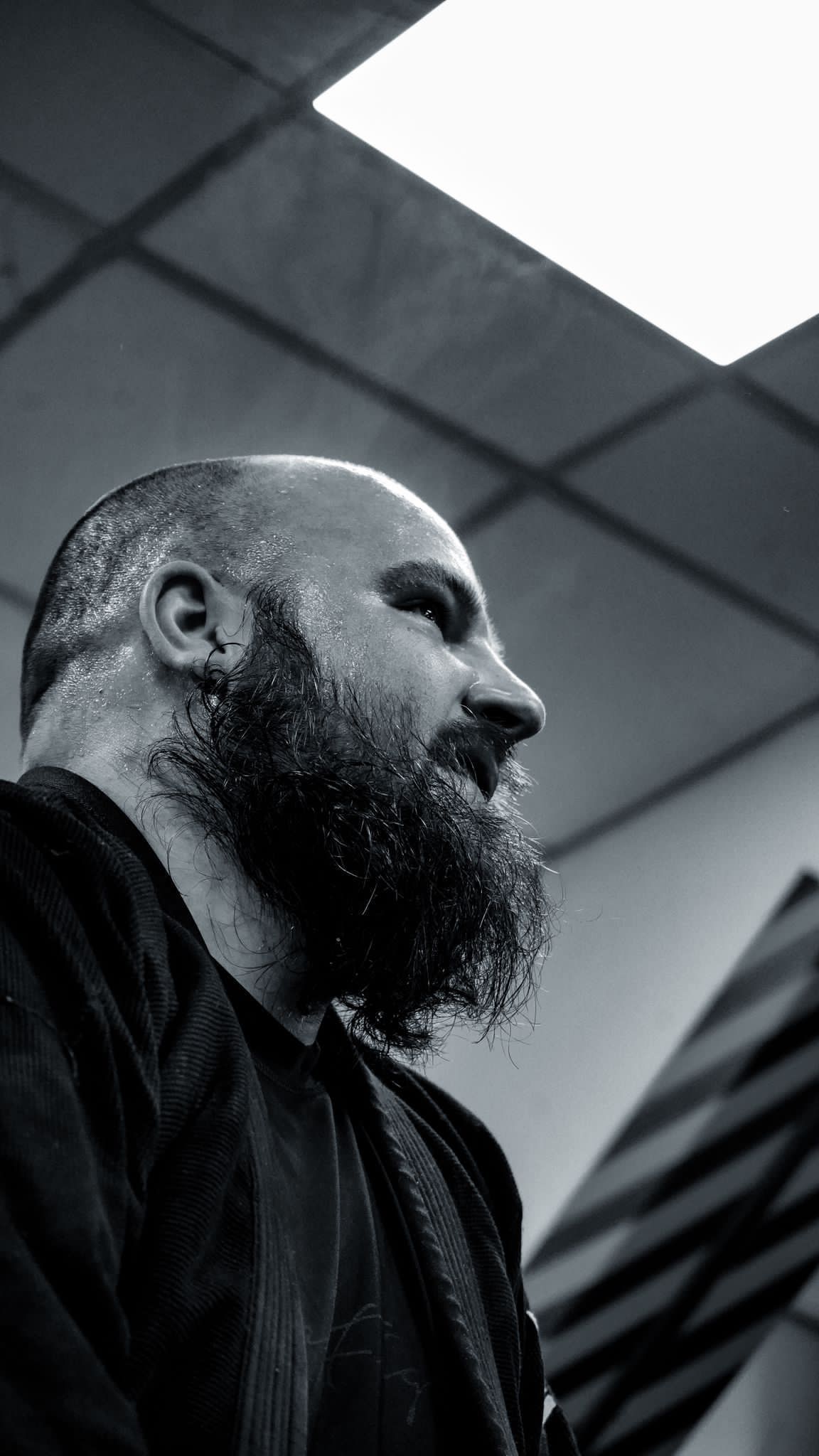 Bearded man in profile under a ceiling light, with a dark jacket and industrial background