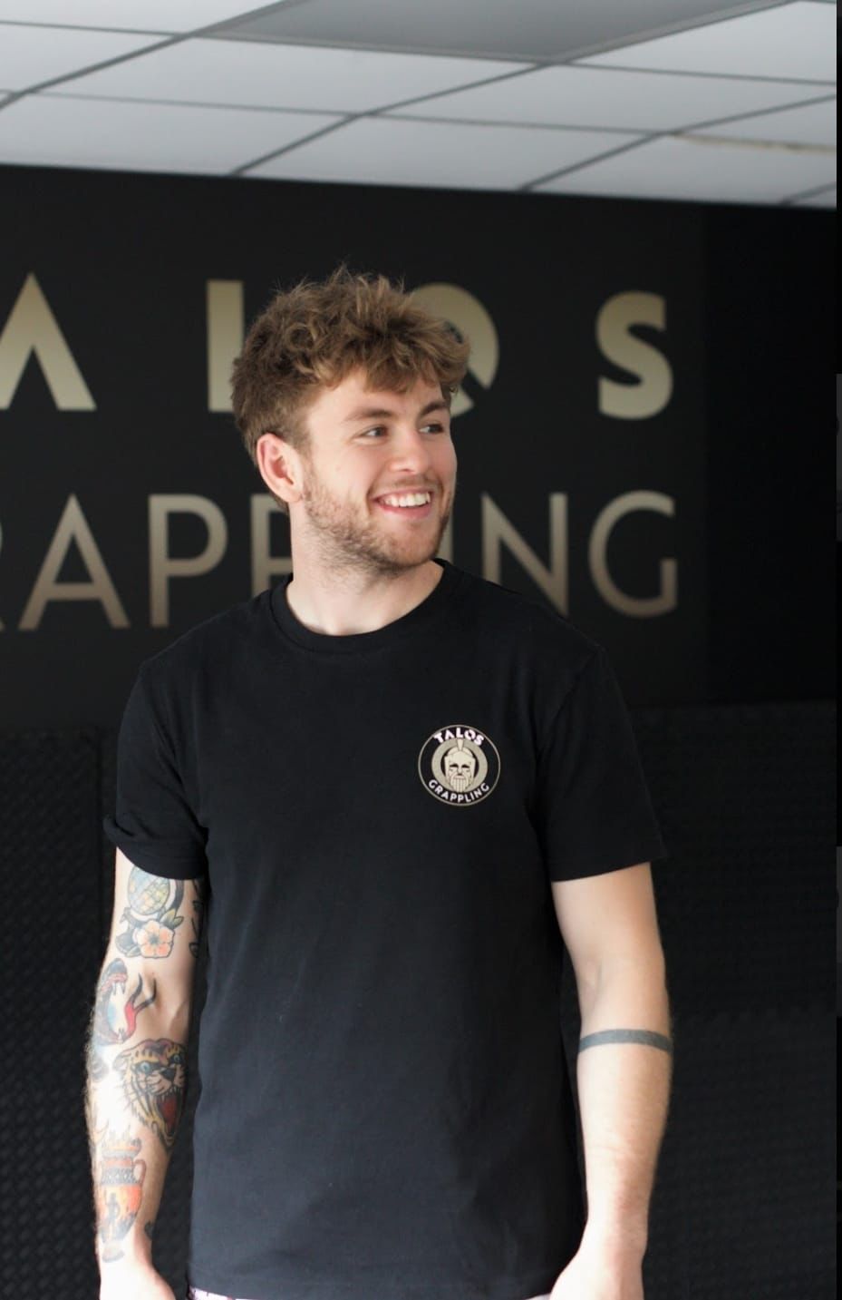 Smiling man in a black T-shirt standing indoors in front of a black wall with large lettering