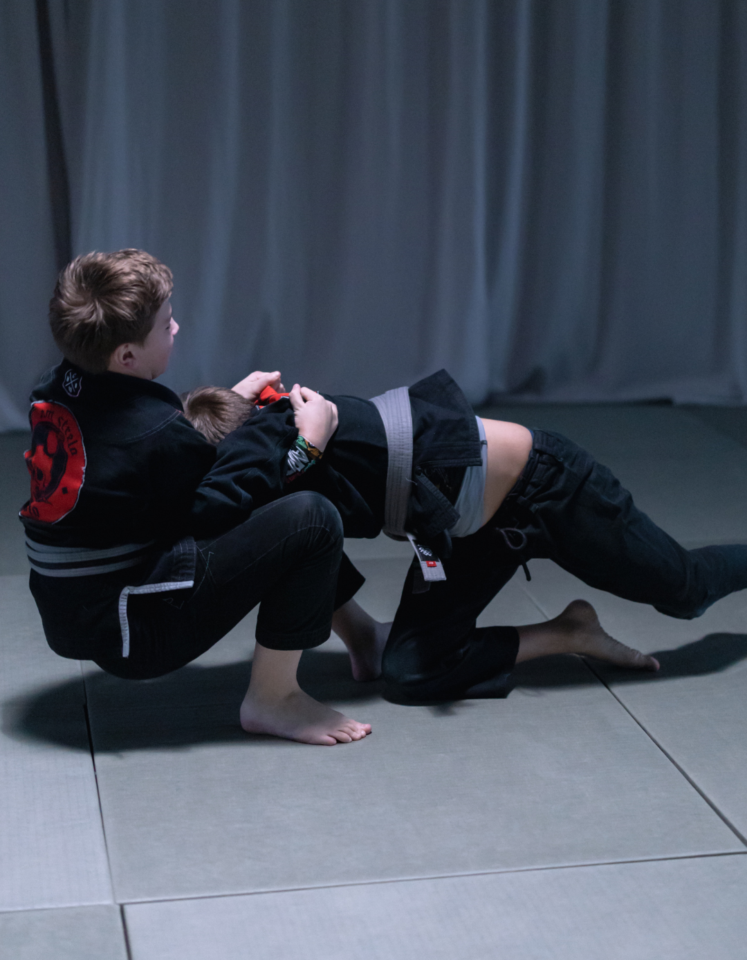 Two martial artists grapple on a mat in a dim training room, one crouched and one bent backward.