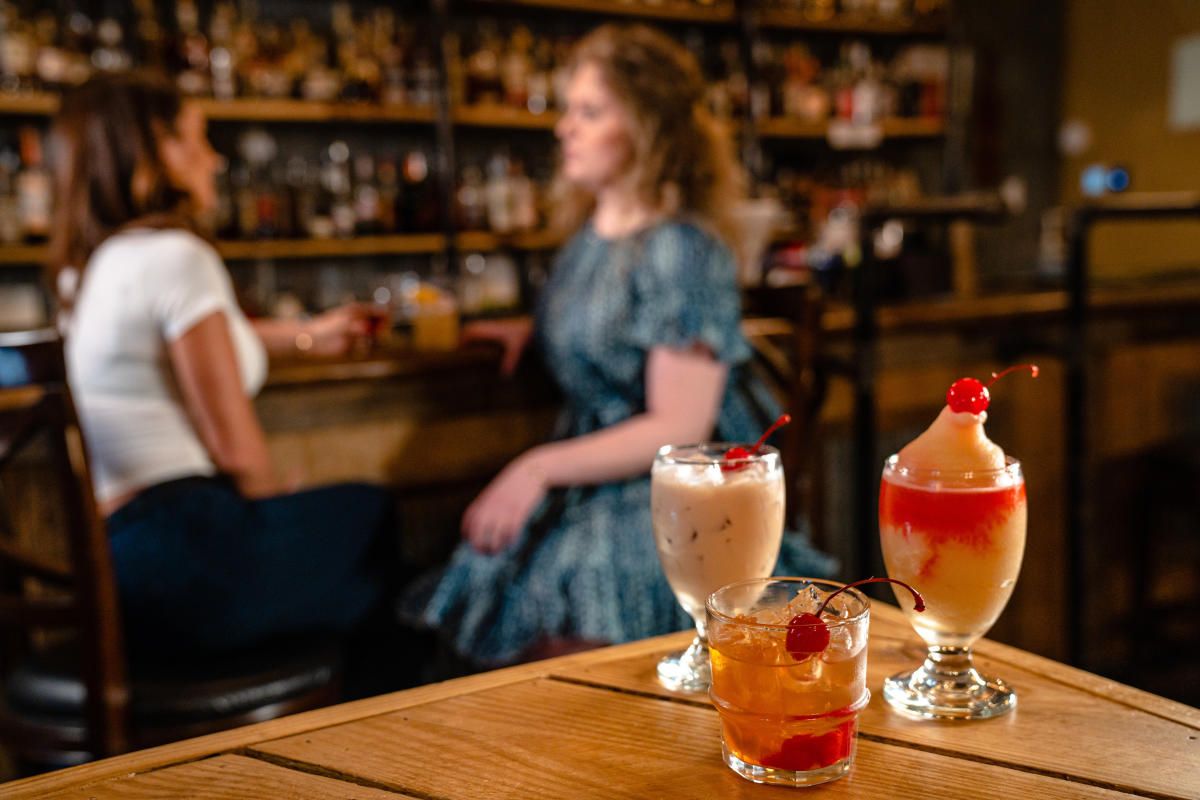 Three cocktails on a wooden table in front of two women sitting at a bar.