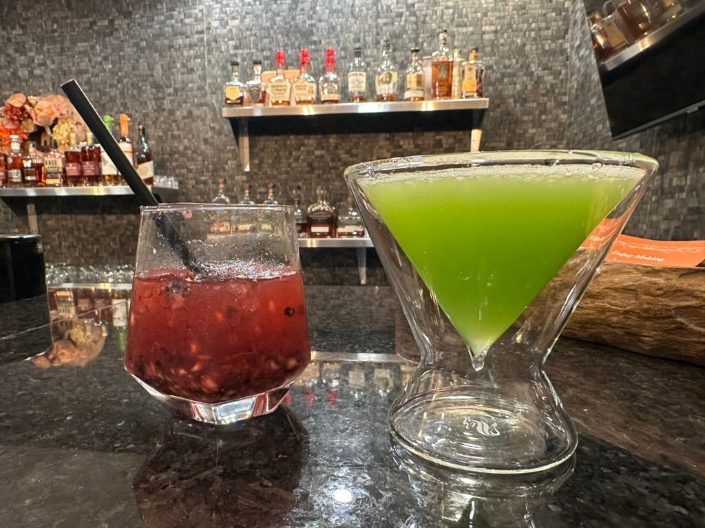Two colorful cocktails, one red and one green, on a bar counter with bottles in the background
