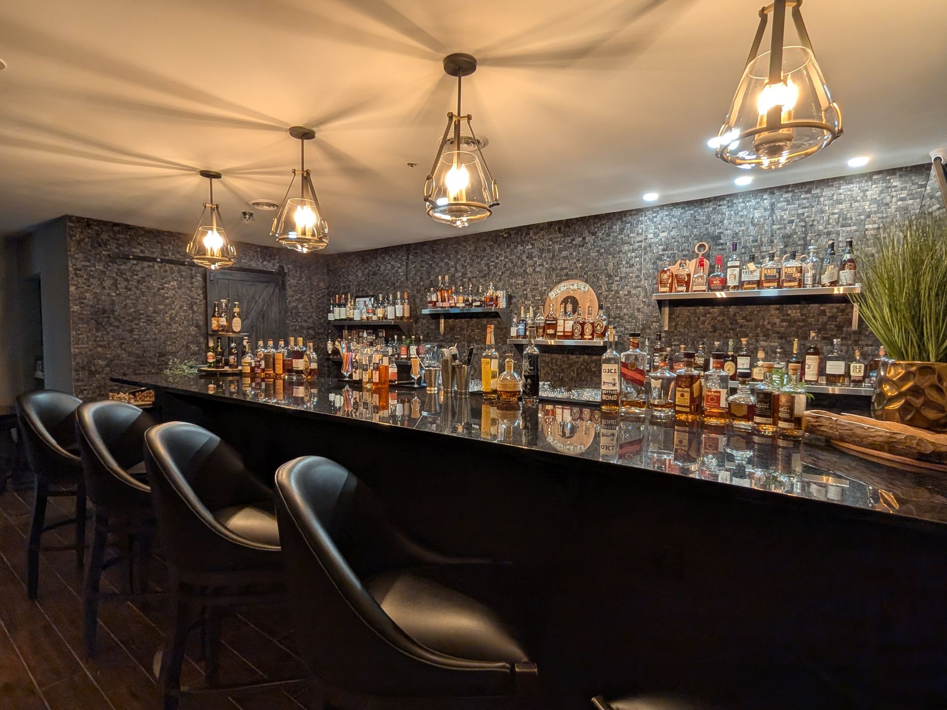 Elegant bar interior with hanging pendant lights, rows of bottles, and black leather stools by a glossy counter