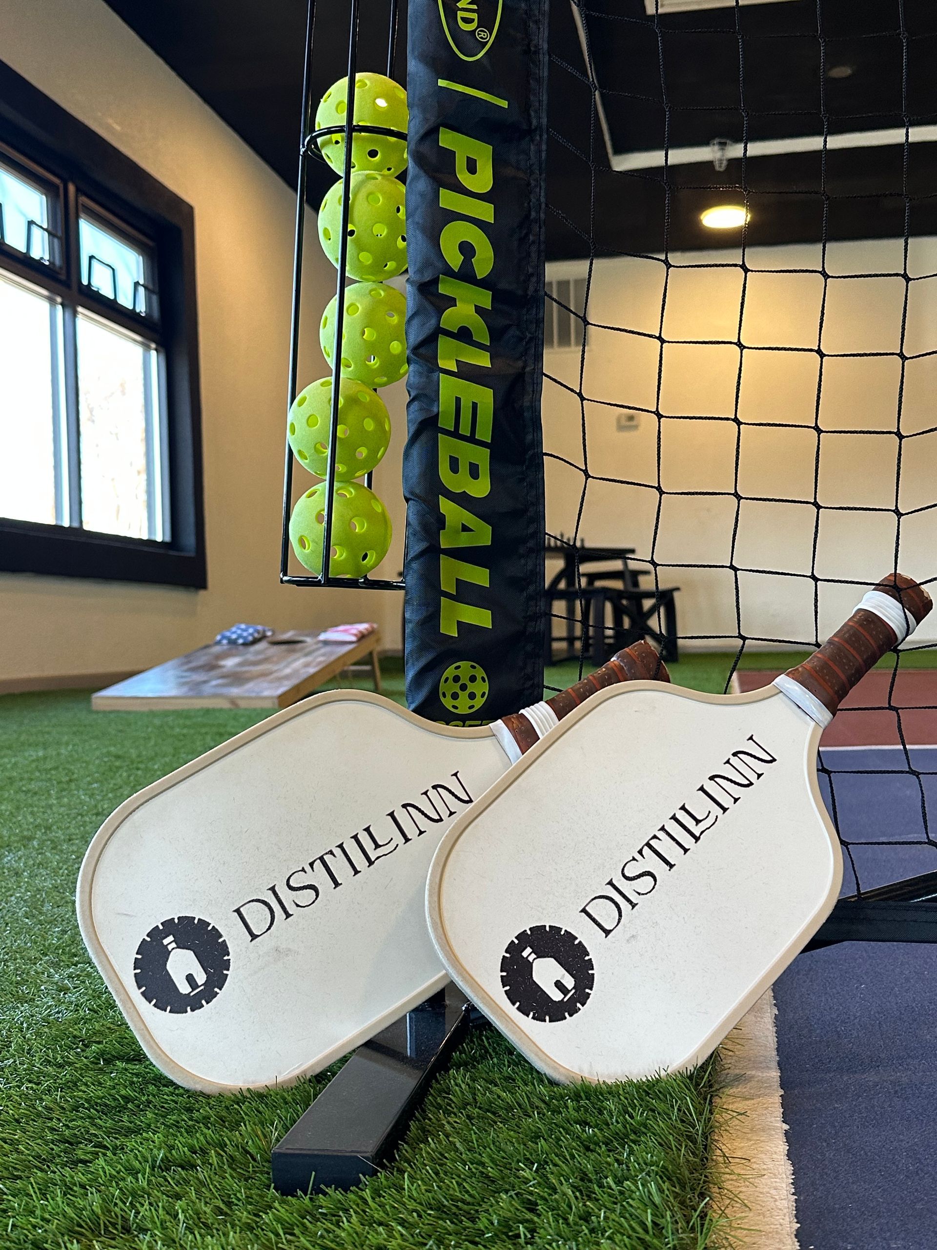 Two pickleball paddles leaning against a net pole, with green balls and an indoor court backdrop.