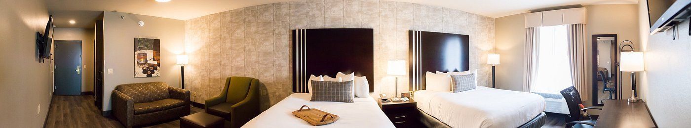 Hotel room with two beds, warm lighting, desk, and wall art across a long interior space