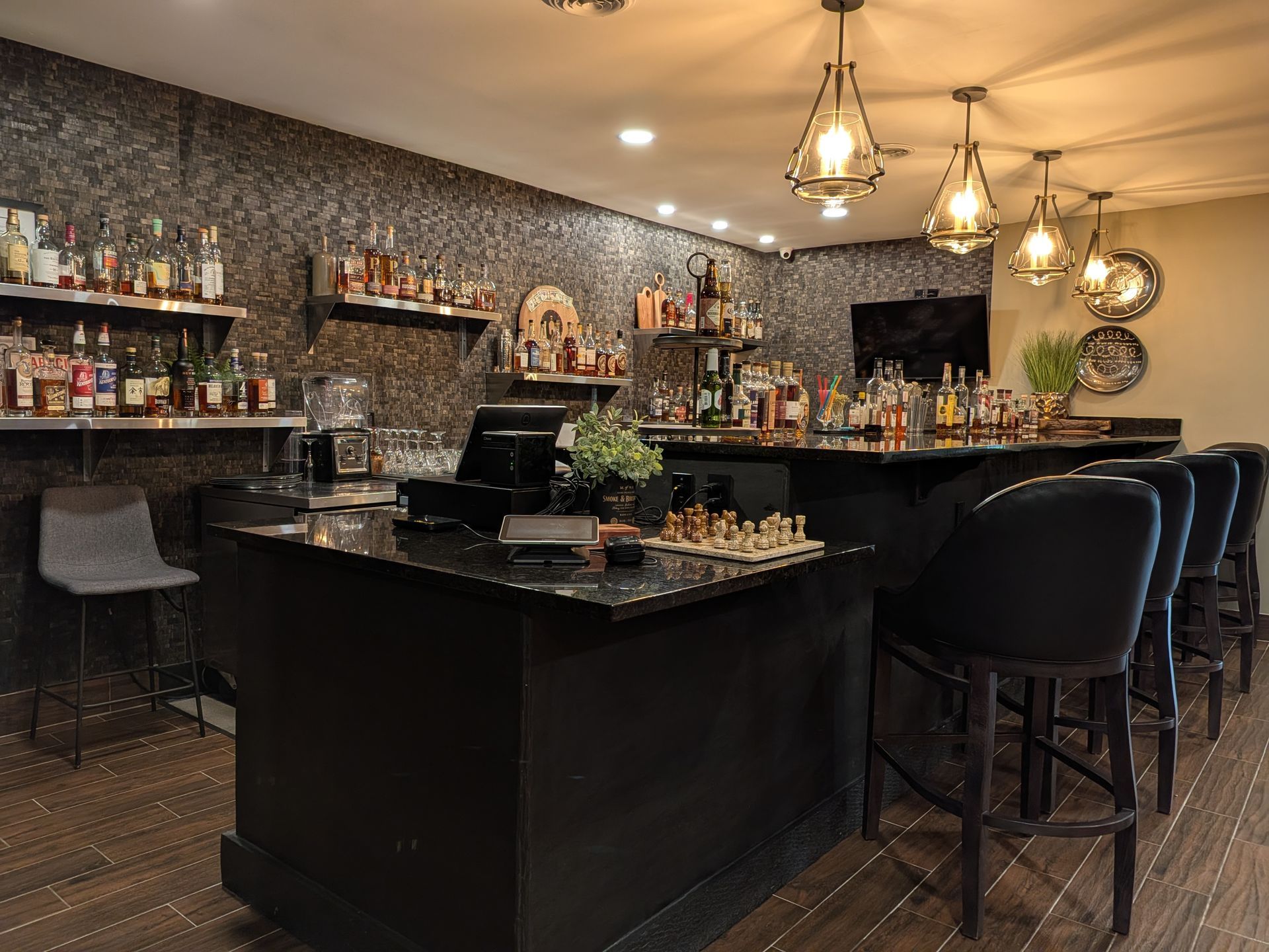 Empty bar with dark counters, black stools, hanging pendant lights, and shelves of bottles.