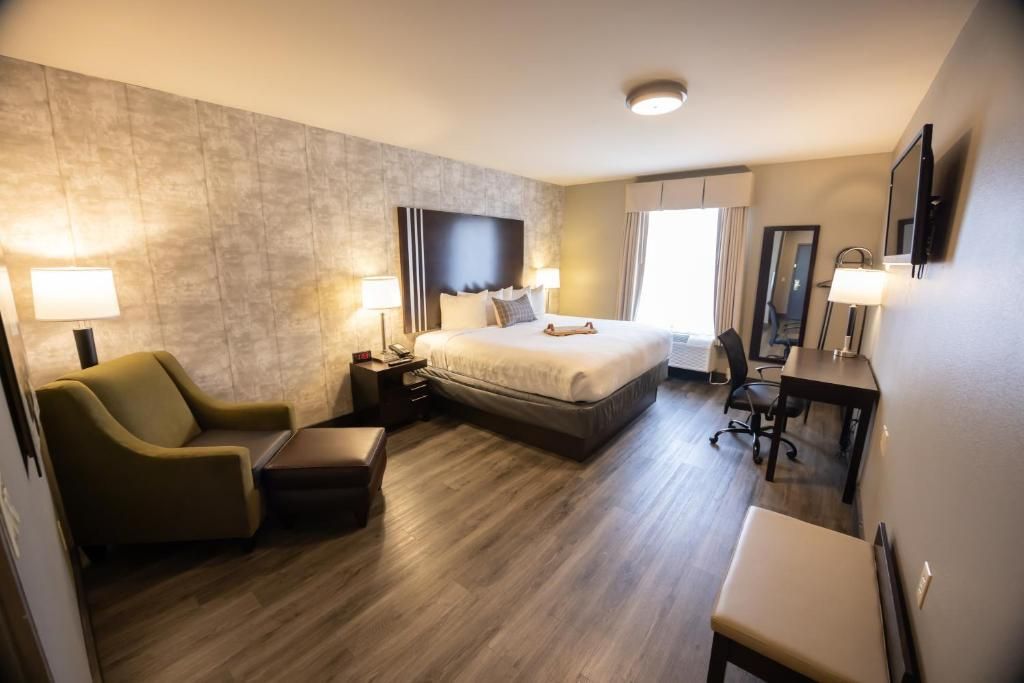 Hotel room with king bed, armchair, desk, TV, and bright window on dark wood floor.