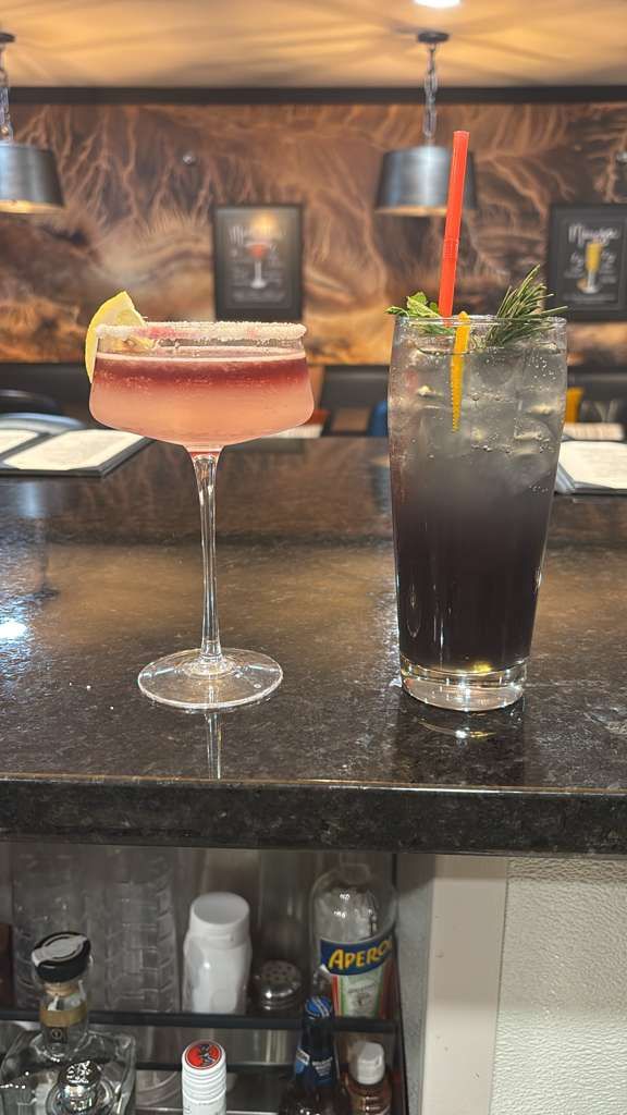 Two cocktails on a bar counter: a pink martini and a dark highball with orange garnish.