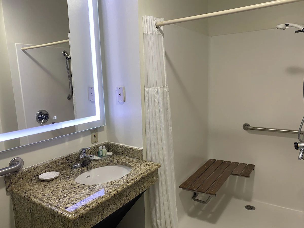 Bathroom vanity with granite sink counter and mirror beside a shower with bench and curtain.
