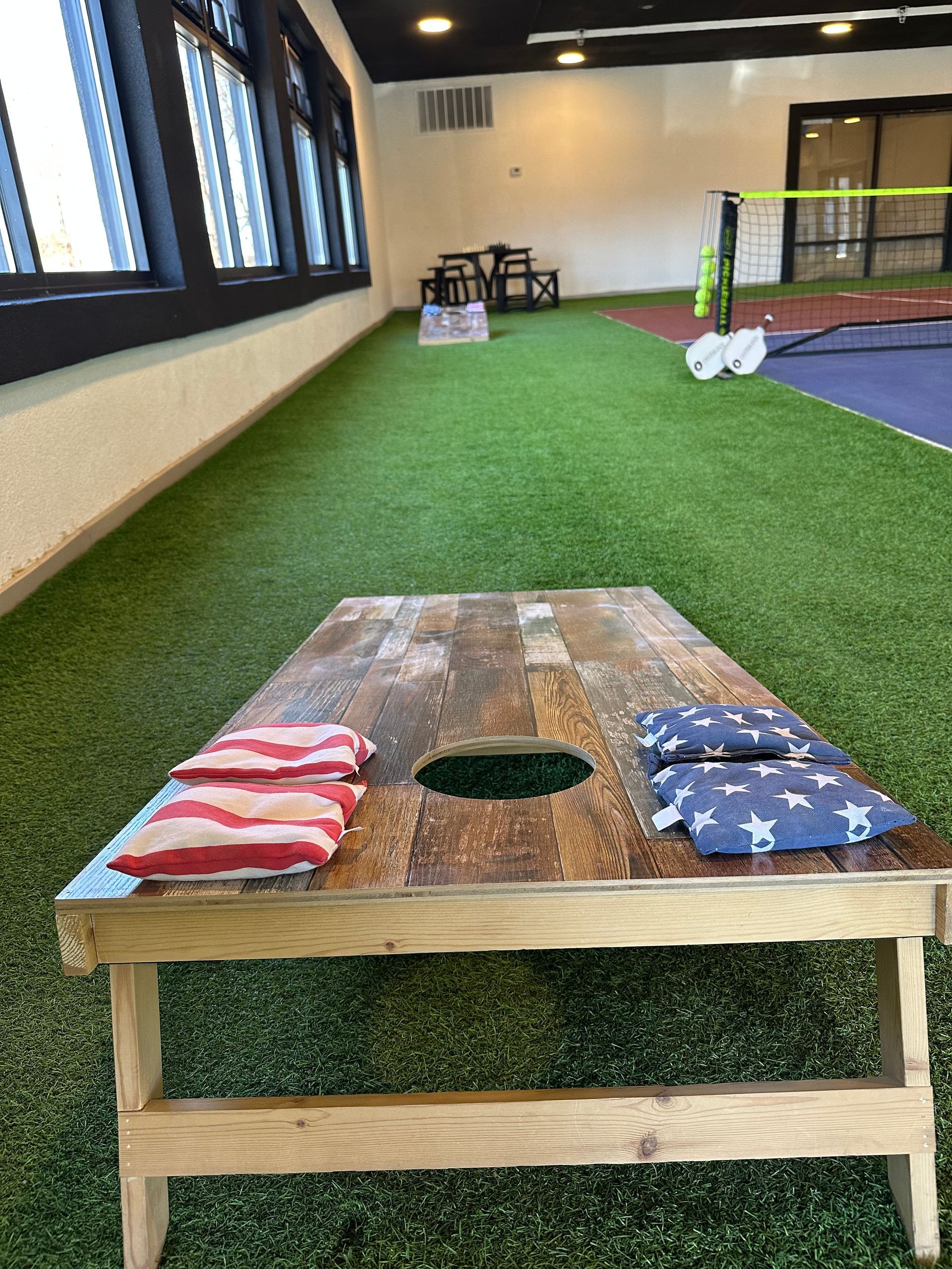 Cornhole board on indoor turf beside a trampoline, with red and blue bean bags on top.