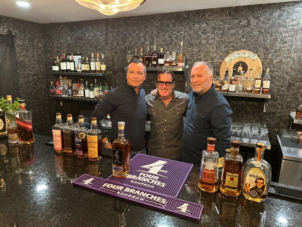 Three people behind a bar with liquor bottles and a purple mat, posing in a dimly lit lounge.