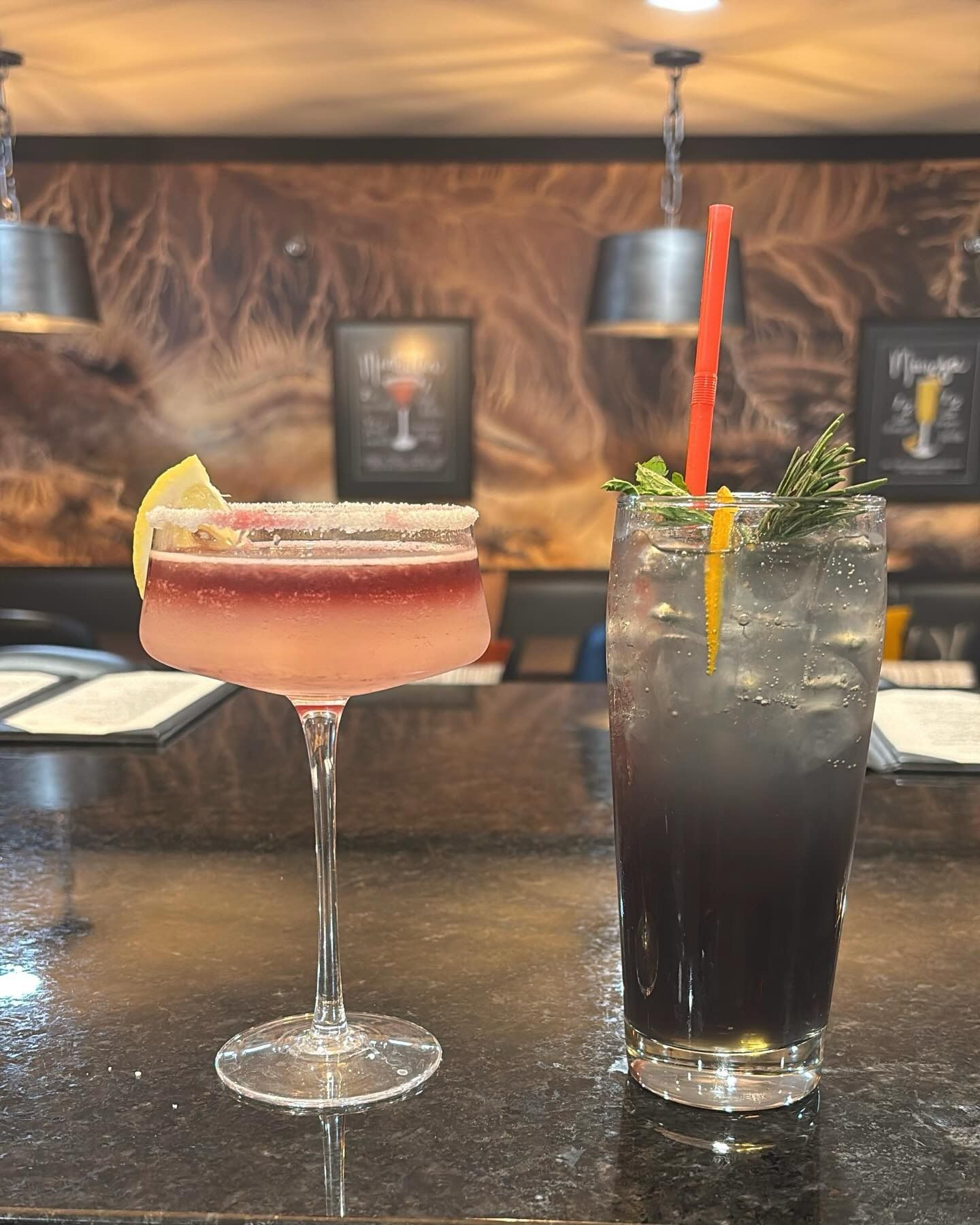 Two cocktails on a bar counter: a pink margarita and a dark highball with herbs and citrus garnish.
