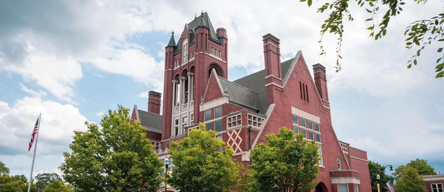 Red brick Gothic-style building with tall towers, trees in front, and a partly cloudy sky