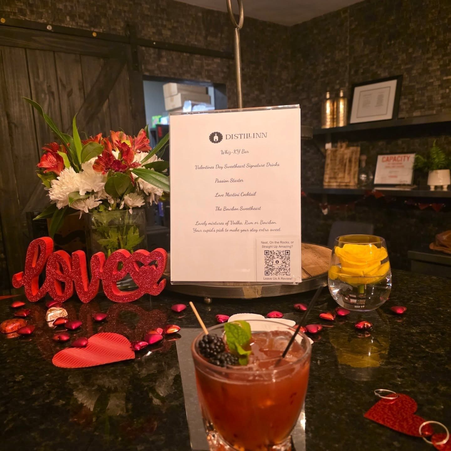 Romantic cocktail table with red decorations, flowers, menu, and two drinks on a dark bar top