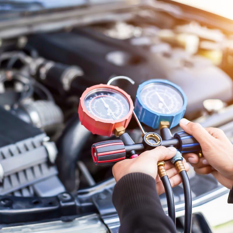 Hands holding HVAC manifold gauges near a car engine, with red and blue dials connected to hoses