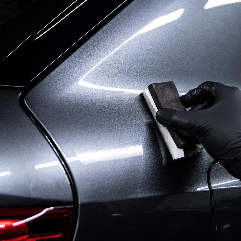 Gloved hand polishing a gray car’s rear panel with a sponge in a close-up detail shot