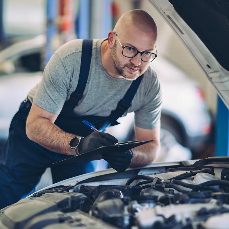 Mechanic in gray shirt and apron inspecting a car engine with a clipboard in a garage