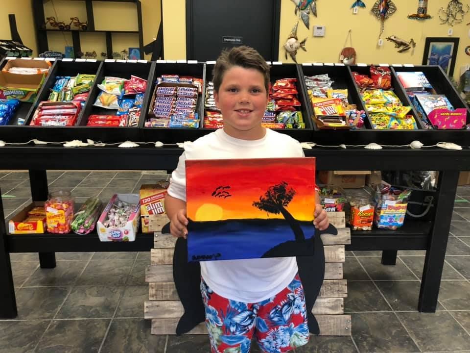 Boy holding painting of a sunset in front of candy display in a USA shop.
