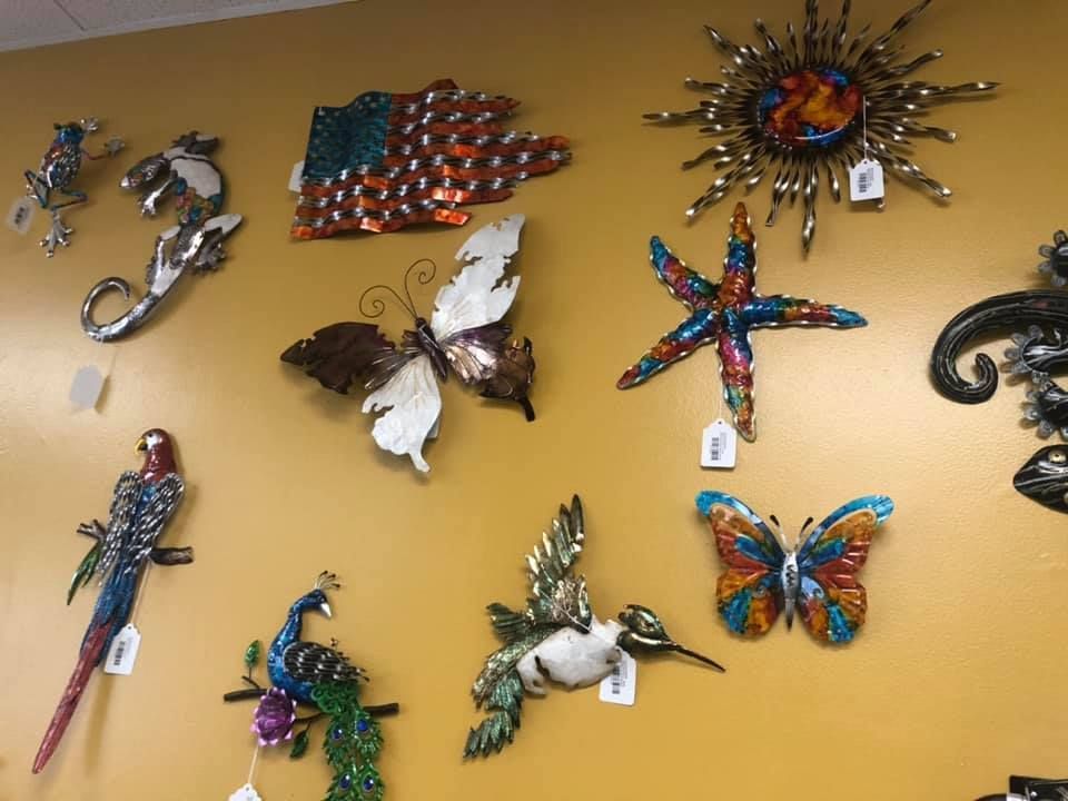 Metal art pieces including a parrot, butterflies, and a sunburst, displayed on a yellow wall in a USA shop.