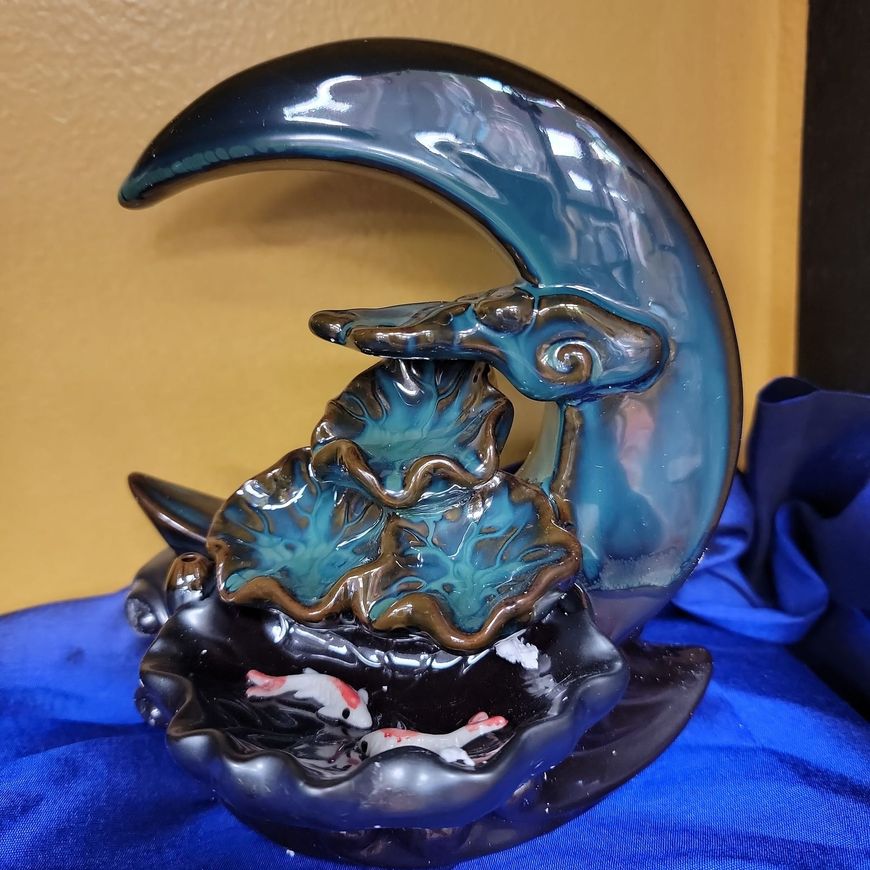 Blue ceramic moon-shaped incense burner with cascading lotus leaves and koi fish, resting on a blue fabric surface.