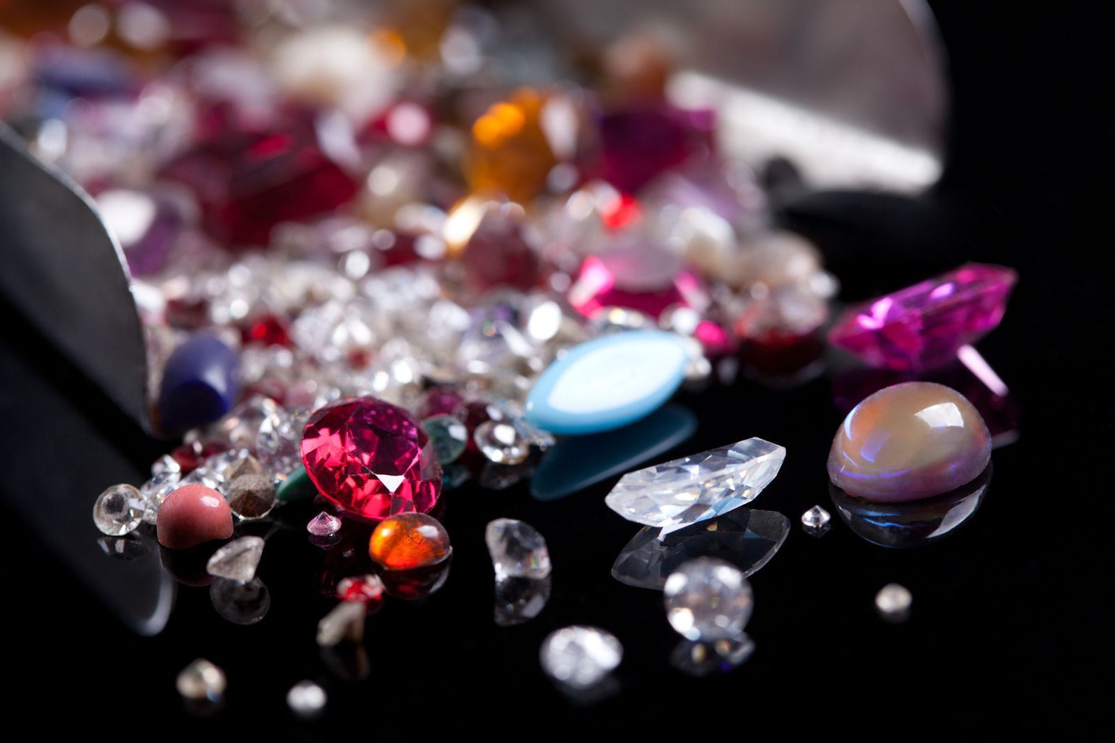 Assorted colorful gemstones spilled on a shiny black surface with a silver scoop.
