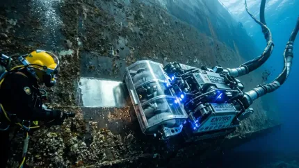 Diver in yellow helmet inspecting a blue underwater robot against a rocky seafloor