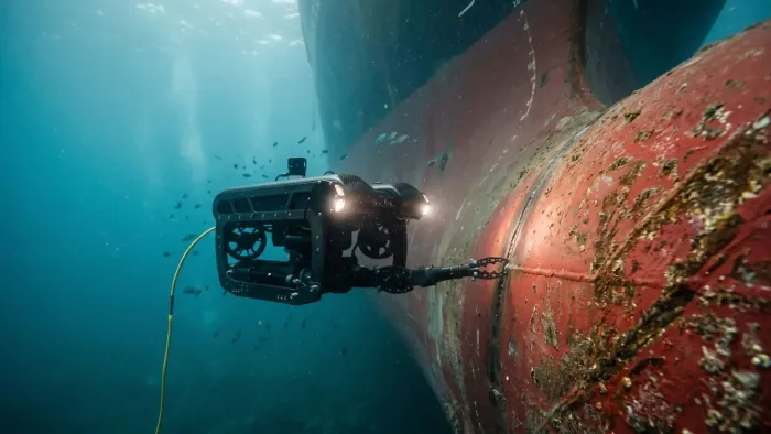 Underwater camera inspecting a large ship hull, with red and black equipment in blue water