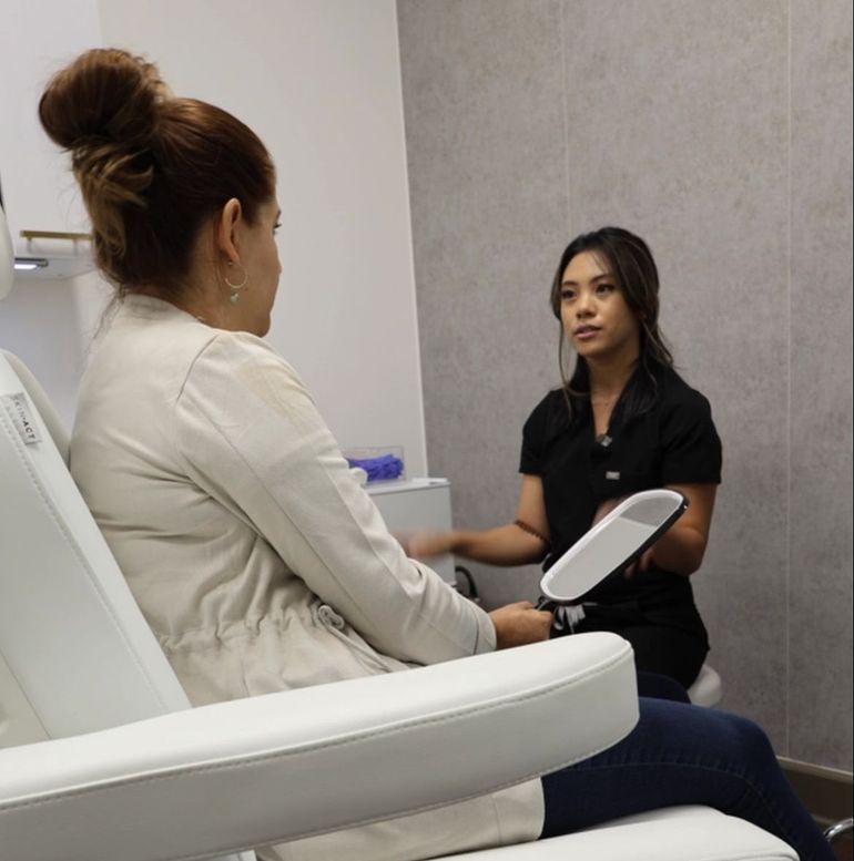 Two women in a clinic consultation room, one seated with a clipboard, the other speaking from a chair.