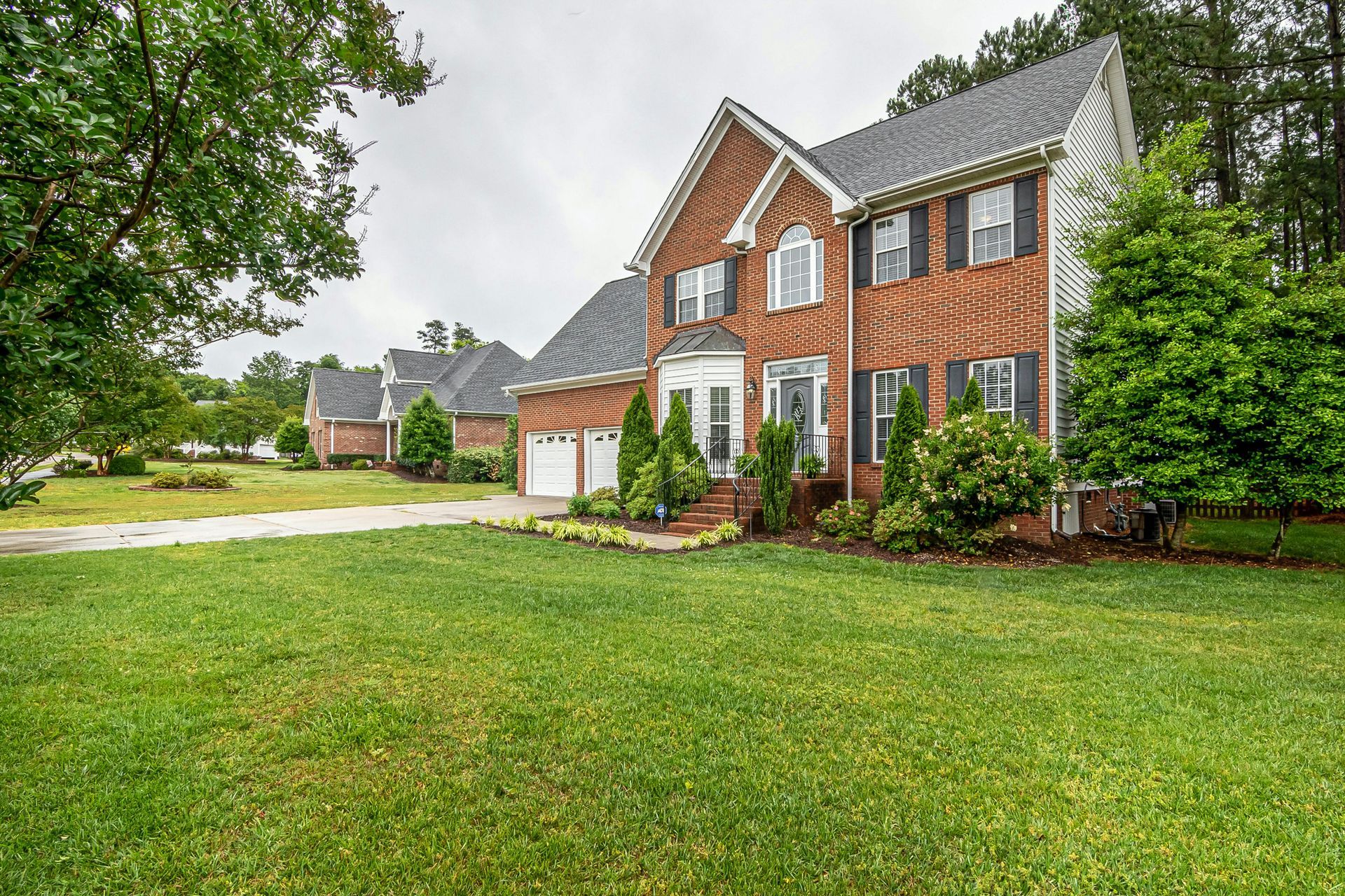 Red brick two-story house with white trim, a garage, and a large green lawn under overcast skies
