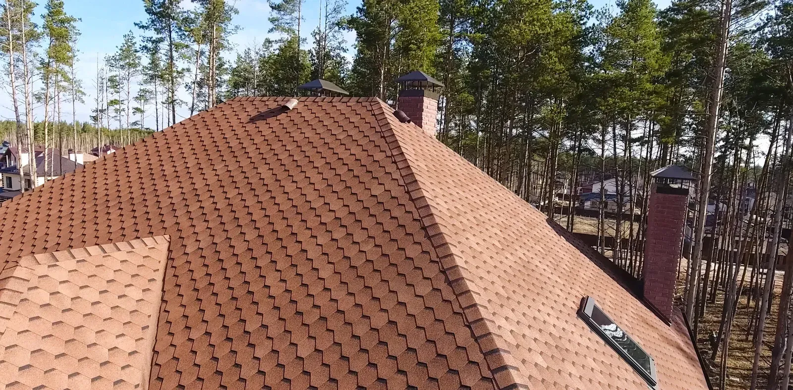 Brown shingled roof with two chimneys, surrounded by trees under a clear sky