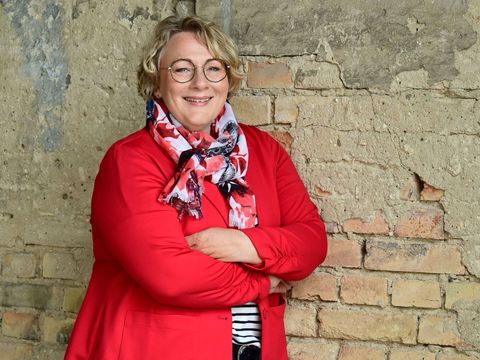 Birgit Hasse in einer roten Jacke und einem Schal steht vor einer Backsteinmauer und lächelt in die Kamera.