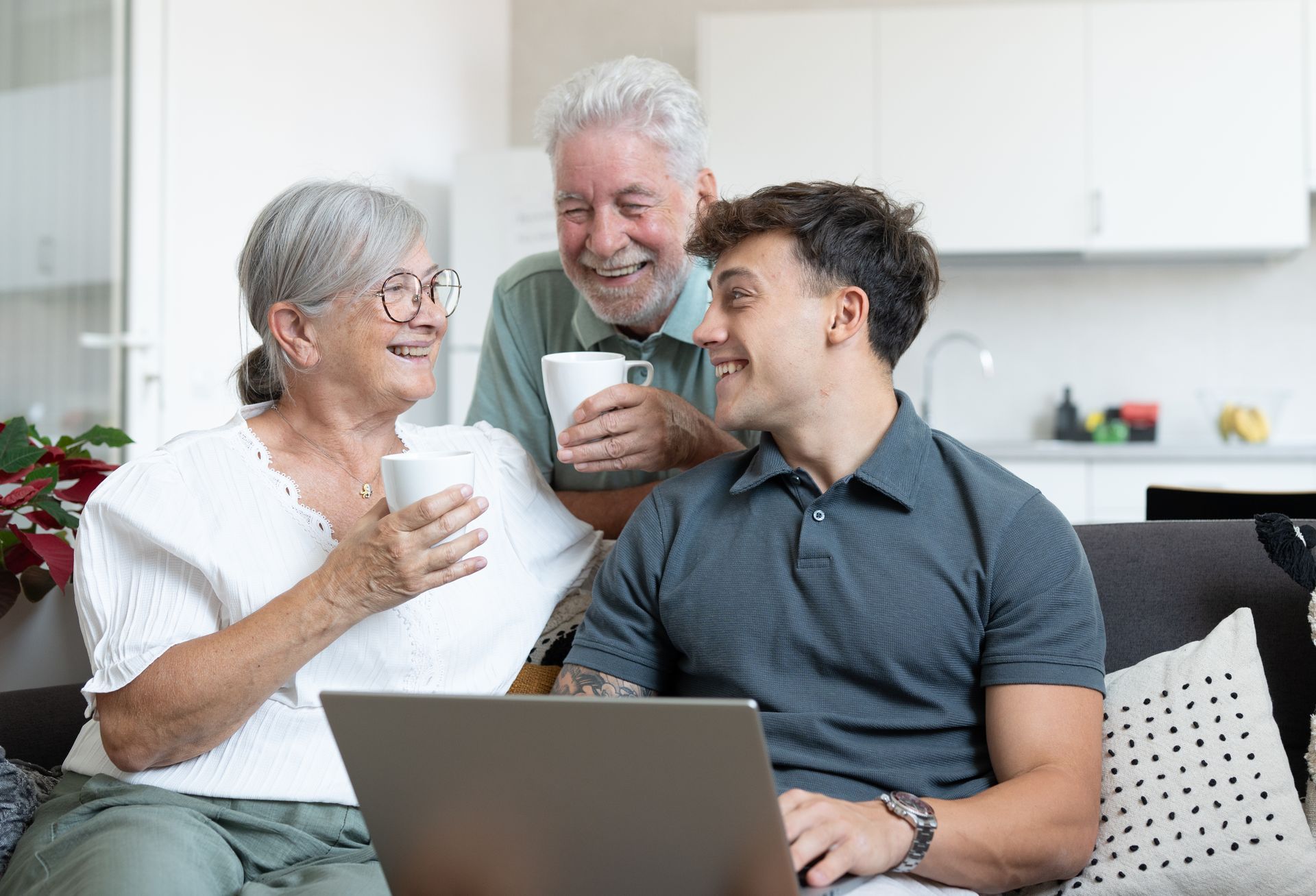 Three smiling people on a couch sharing drinks and looking at a laptop in a bright living room