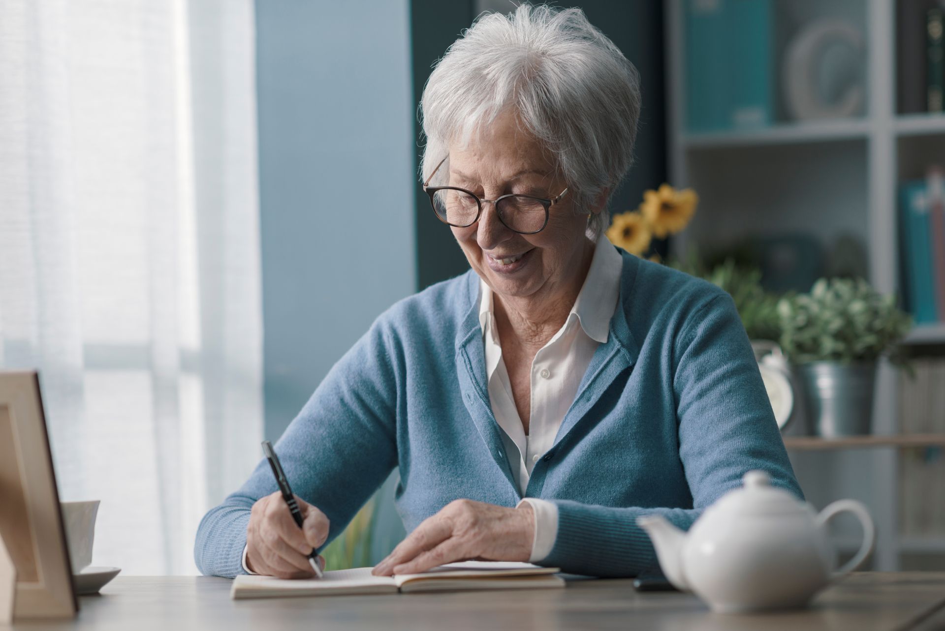 Older woman writing in a notebook at a table in a bright room