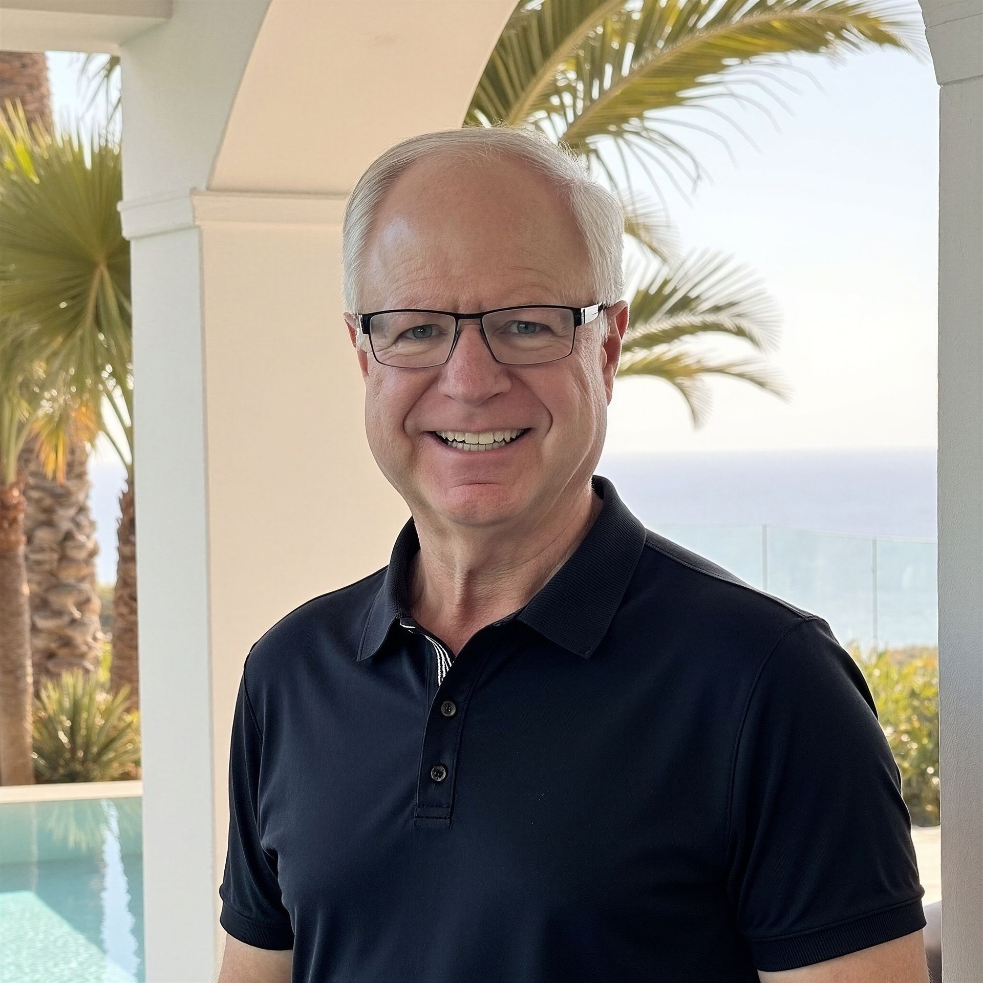 Smiling man in glasses and a black polo stands on a sunny balcony with palm trees and ocean in the background