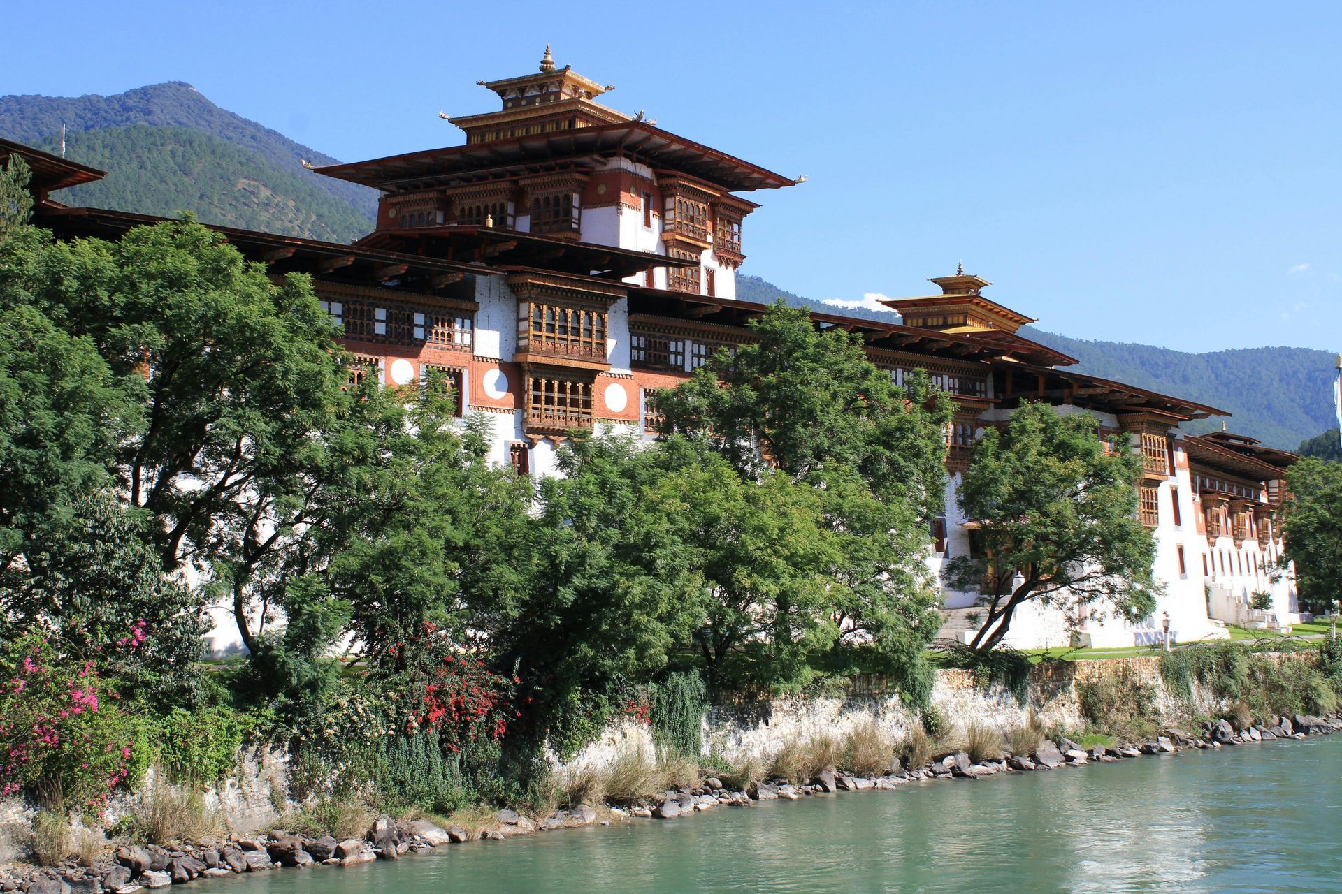 Traditional Bhutanese-style building beside a river, with trees and mountains under a clear blue sky