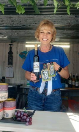 Smiling woman in blue shirt holding a wine bottle and gift basket at a market stall