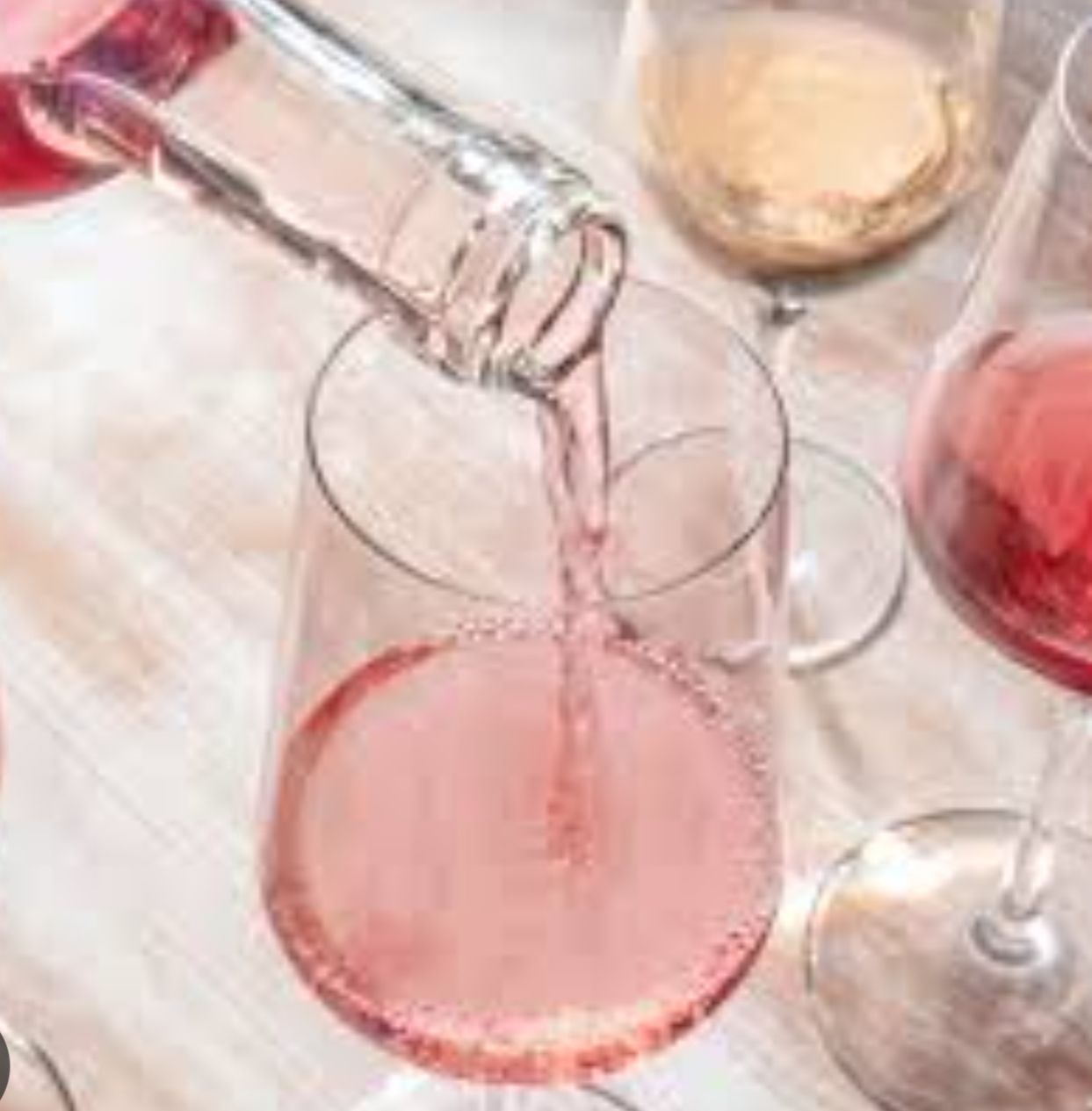 Clear wine being poured into a glass, with a pink drink and glasses on a table.