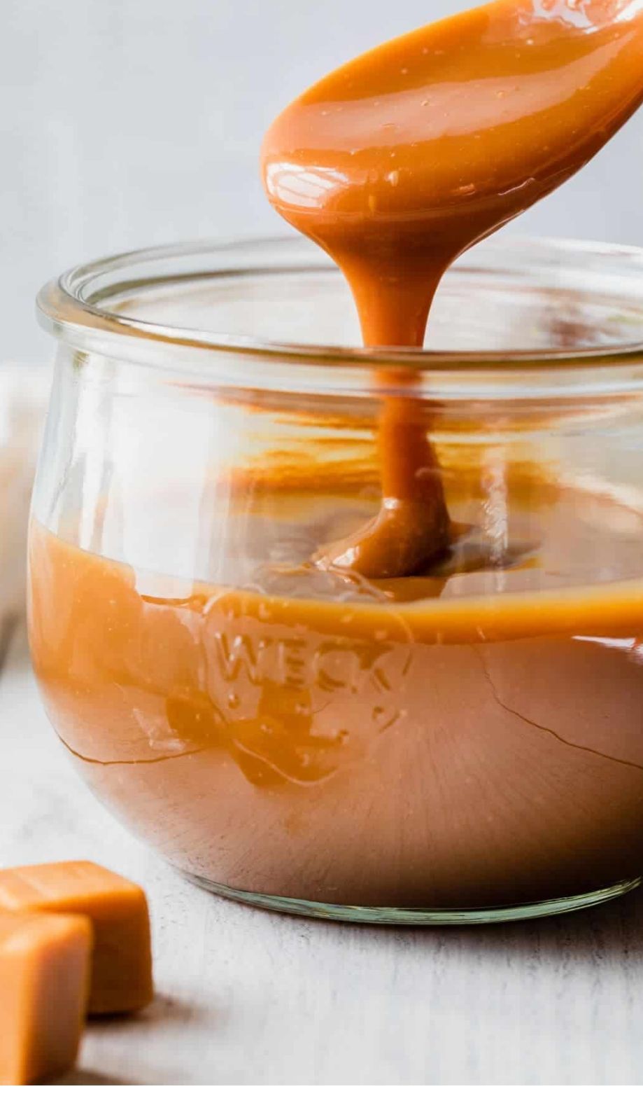 Spoon lifting caramel sauce into a glass jar, with more sauce beside it on a light surface